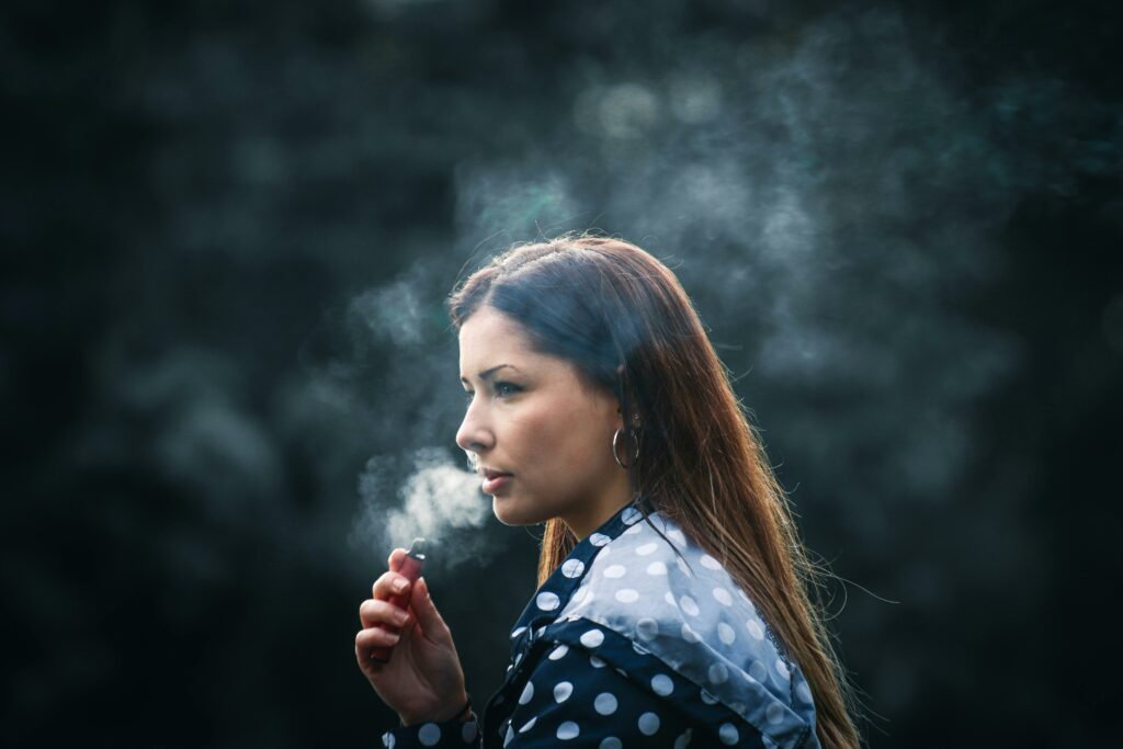 Mulher loira de cabelos lisos em meio à fumaça de um cigarro eletrônico que ela está assoprando